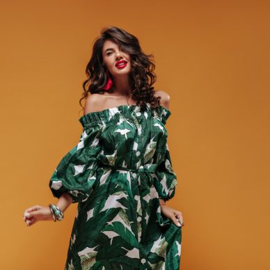 Charming wavy haired woman with bright lipstick in green modern dress and accessories looking into camera and posing on isolated backdrop..