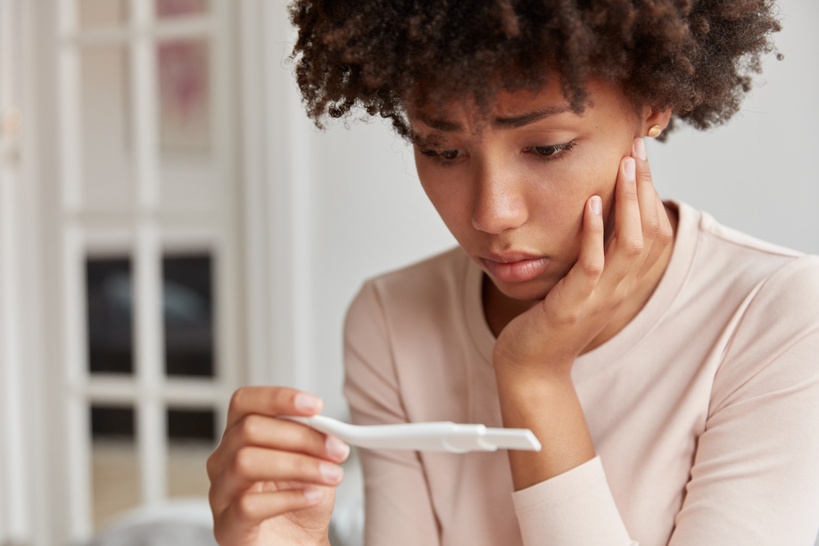 Displeased frustrated African American woman looks stressfully at pregnancy test, finds out she will become mother, looks desperately at result. Close up shot of young lady has problems with fertility