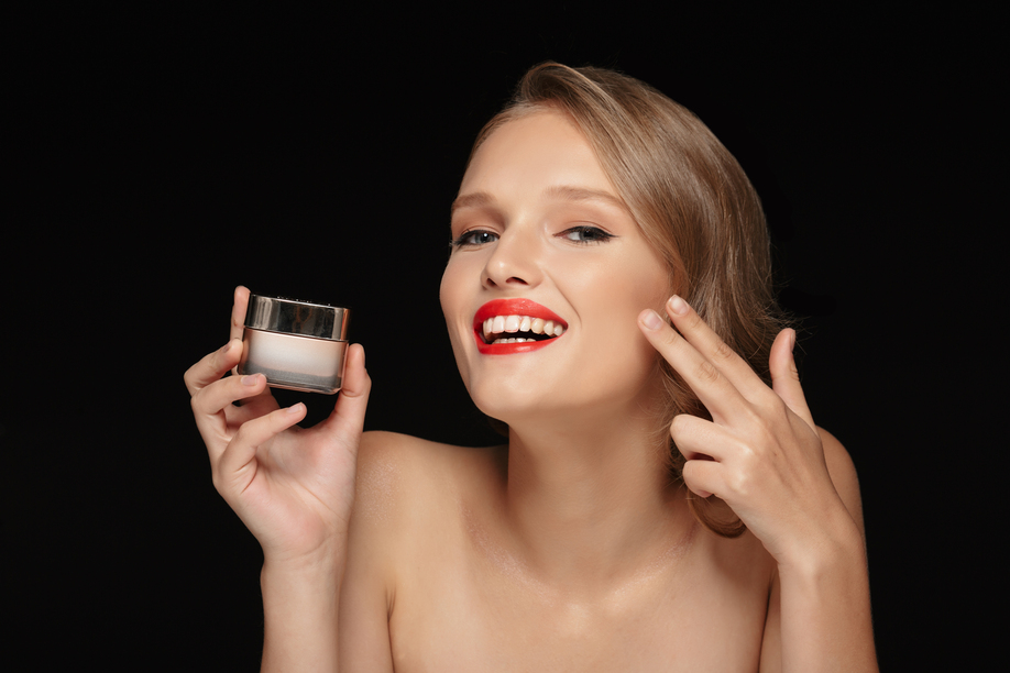 Portrait of young joyful woman with wavy hair and red lips holding beauty cream in hand happily looking in camera over black background
