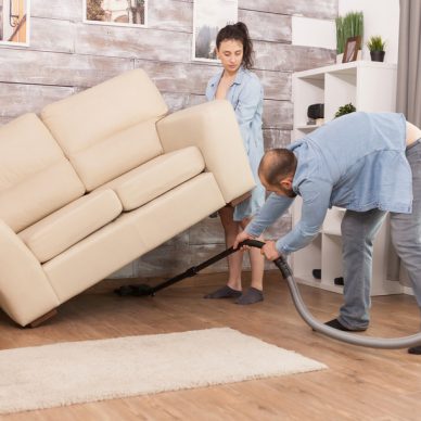 Wife picks up sofa while her husband is cleaning the dust under it with vacuum cleaner.