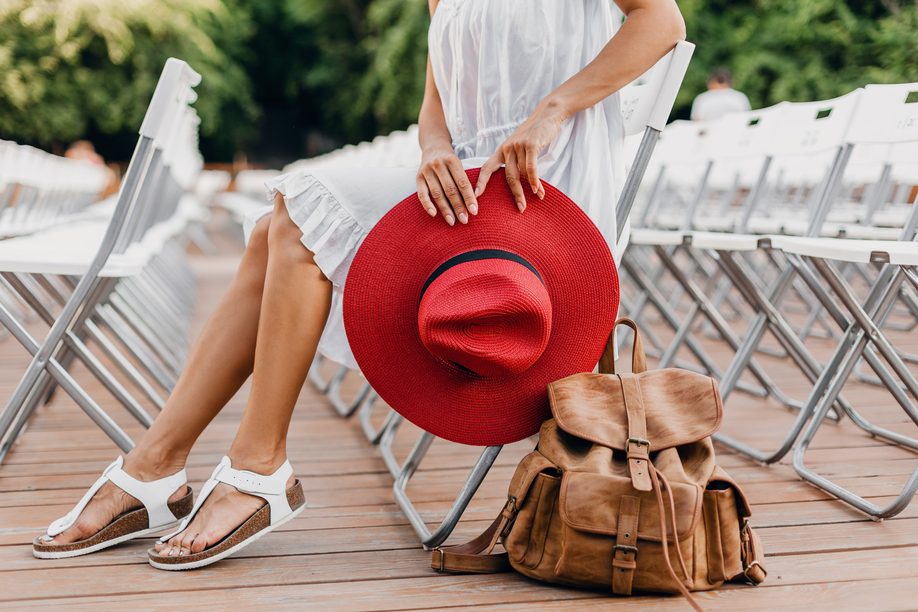 close-up details of woman in white dress, red hat sitting in summer open air theatre on chair alone, spring street style fashion trend, accessories, traveling with backpack, skinny legs in sandals