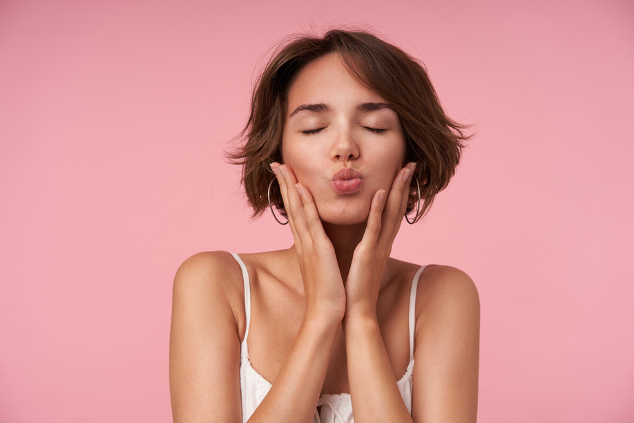Close-up of pleasant looking brunette female with short haircut posing over pink background with eyes closed, holding her face with raised hands and folding lips in kiss