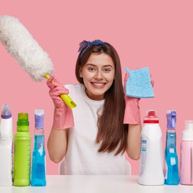 Joyful dark haired woman carries rag and brush, smiles happily, dressed in casual clothes, sits at white desk with cleaning products, isolated over pink background. Happiness, housekeeping concept