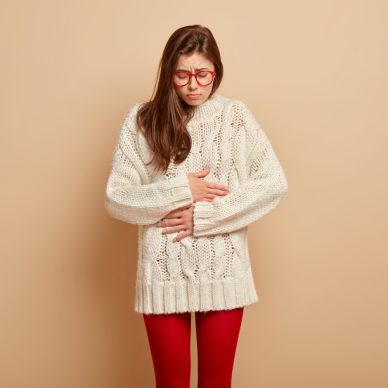 Dissatisfied woman suffers from bellyache, keeps hands on stomach, feels cramps, being very hungry, has sad expression, dressed in oversized white jumper and red tights. People, diarrhea, dieting