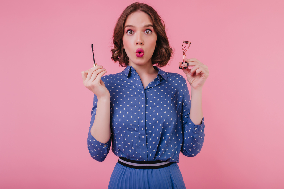 Shocked dark-eyed girl in trendy blouse posing on pink background with mascara. Indoor photo of brunette surprised young woman doing her eyelashes.