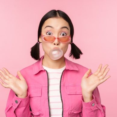 Stylish asian girl blowing bubblegum bubble, chewing gum, wearing sunglasses, posing against pink background. Copy space