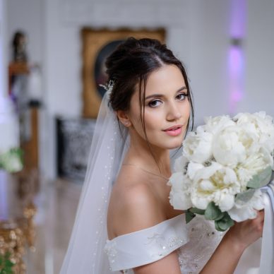 Beautiful brunette caucasian bride is holding bouquet of white peonies and looking straight indoor