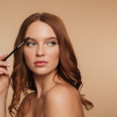 Beauty portrait of calm ginger woman with long hair looking away and posing sideways while combing her eyebrows with brush over cream background