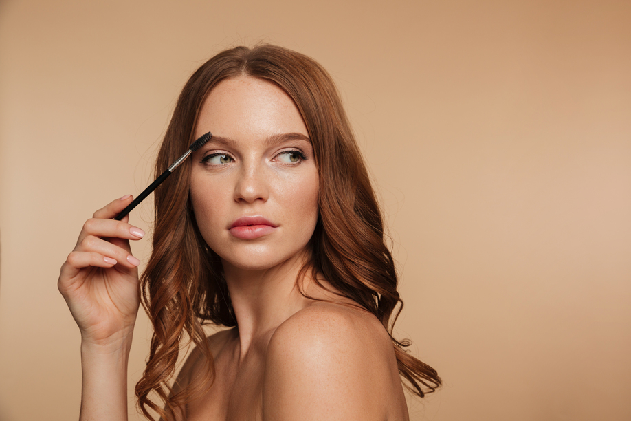 Beauty portrait of calm ginger woman with long hair looking away and posing sideways while combing her eyebrows with brush over cream background