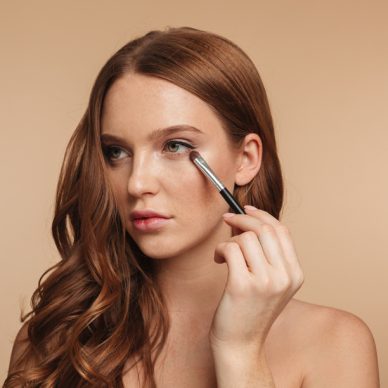 Beauty portrait of mystery smiling ginger woman with long hair looking away while applying cosmetics with brush for eyeshadow over cream background