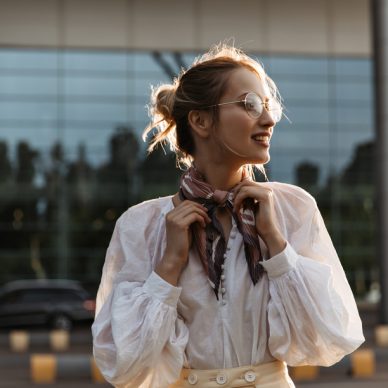 Blonde woman touches her silk brown scarf. Attractive young girl in eyeglasses and white blouse smiles outside.