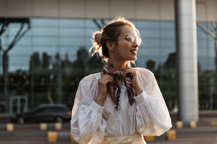 Blonde woman touches her silk brown scarf. Attractive young girl in eyeglasses and white blouse smiles outside.