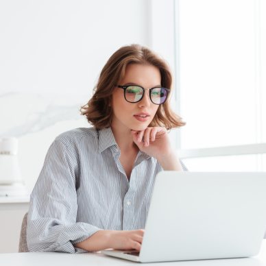 Charming businesswoman in glasses and striped shirt working with laptop computer while siting at home
