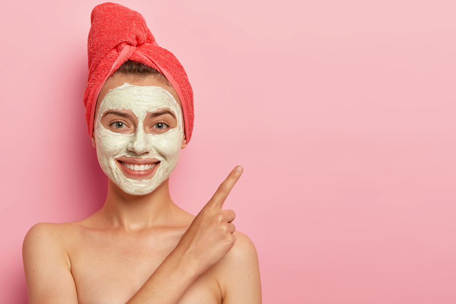 Studio shot of cheerful young woman with gentle smile, points index finger on copy space, advertises beauty item, stands shirtless, wears clay mask and towel poses against pink background. Cosmetology