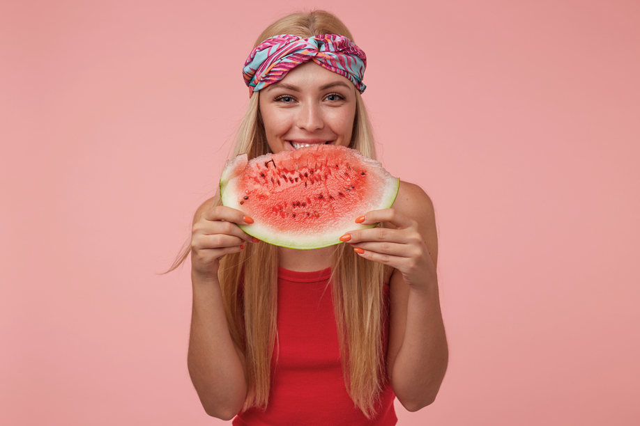 Cheerful young woman with long blond hair standing over pink background, wearing red shirt and colored headband, going to eat watermelon