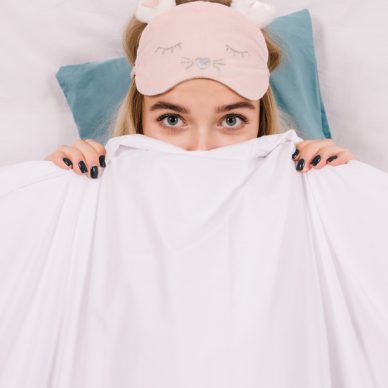 Close-up shot of caucasian girl in sleep mask hiding under blanket. Overhead portrait of refined european woman posing in bed.