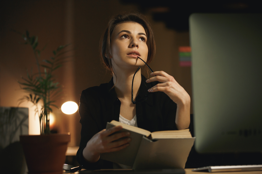 Photo of concentrated young lady designer sitting indoors at night using computer and reading book. Looking aside.