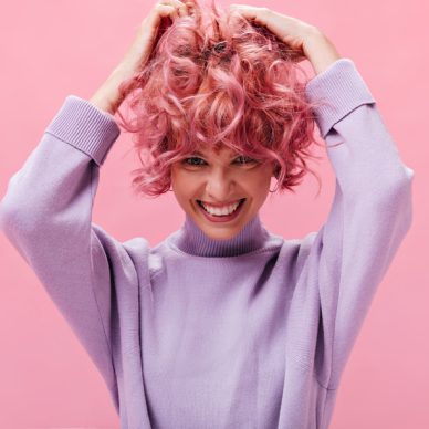 Cute young girl ruffles curly pink hair on isolated background. Pretty short-haired woman in purple sweater smiles sincerely.
