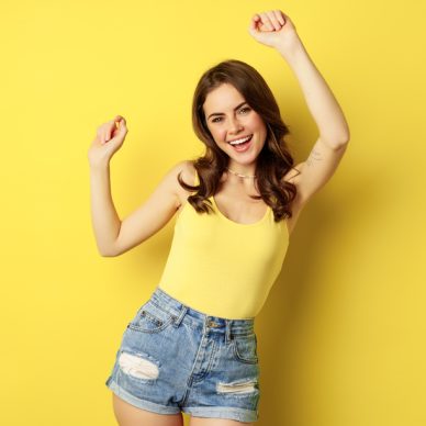 Dancing happy summer girl having fun, raising hands up and rejoicing, standing in tank top and shorts against yellow background.