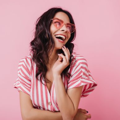 Happy woman with wavy brown hair laughing on pastel background. Jocund girl in striped pink attire smiling in studio