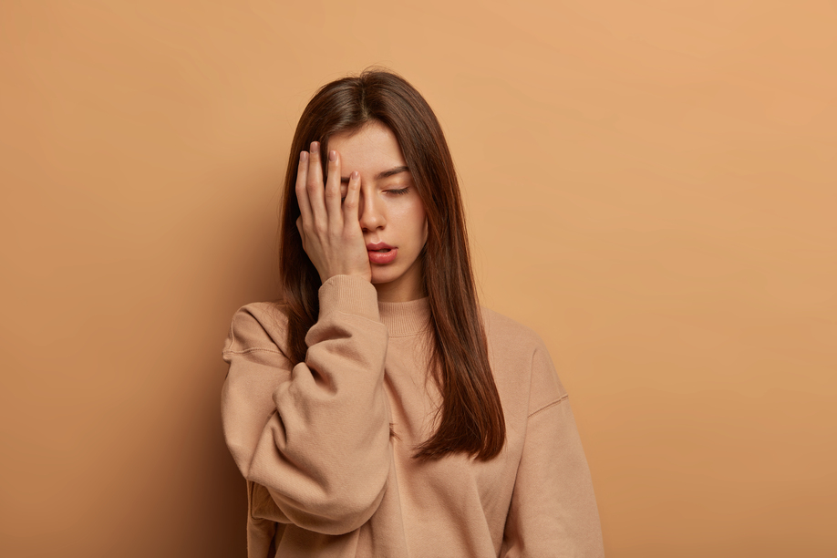 I cannot look at this mess. Frustrated tired woman makes facepalm, stands displeased and uninterested, sighs from tiredness after working long time, wears sweatshirt, isolated on brown wall.