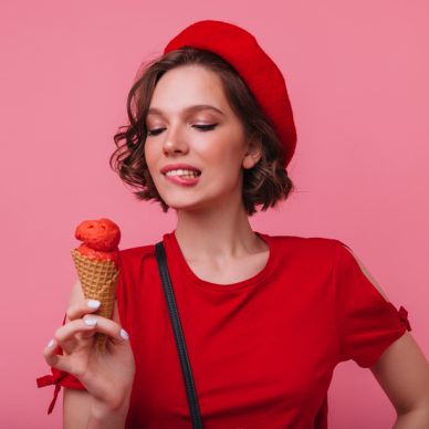 Inspired female model with wavy hairstyle looking at ice cream with smile. Sensual french girl in beret enjoying dessert in studio.