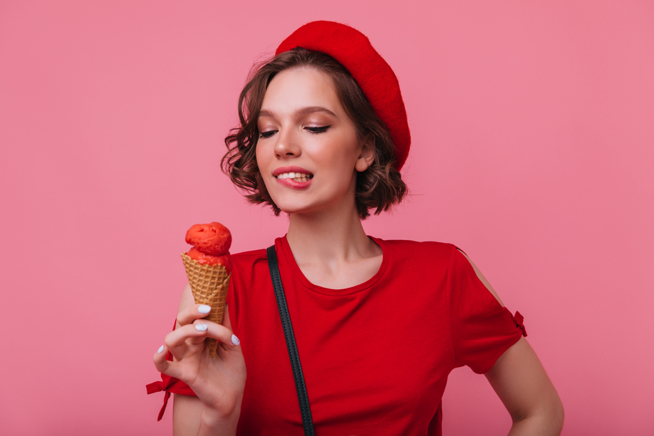 Inspired female model with wavy hairstyle looking at ice cream with smile. Sensual french girl in beret enjoying dessert in studio.