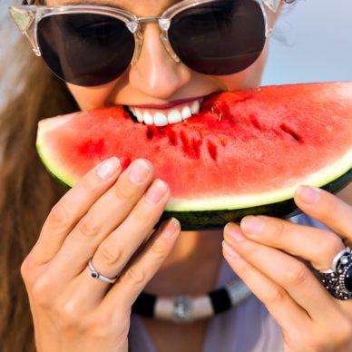 Joyful girl with long light-brown hair biting a watermelon. Close-up portrait of excited female model in big dark sunglasses enjoying favorite food with smile.