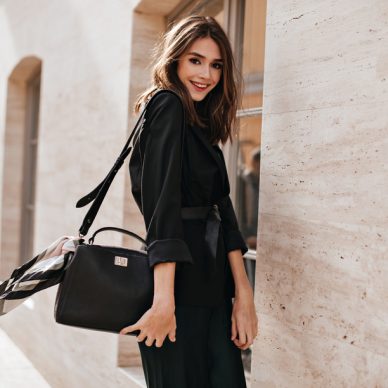 Joyful young brunette with fluffy hair, red lips, trendy dress and black jacket, belt on waist standing in profile at sunny street and smiling against light building background