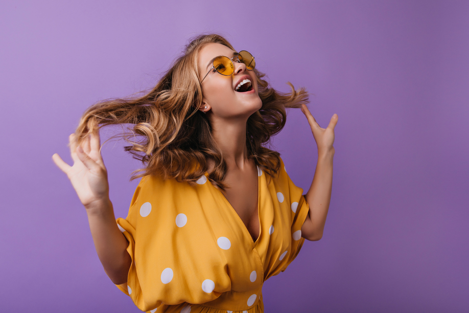Lovable european woman fooling around on violet background. Fashionable tanned girl playing with her blonde wavy hair.