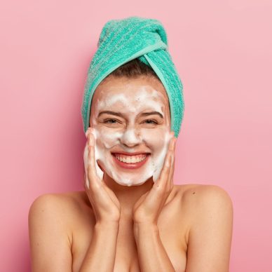 Pampering and hygiene concept. Happy young European woman massages cheeks, aplies bubble foam, washes face, smiles positively, has naked body, enjoys taking shower, wants to have clean skin.