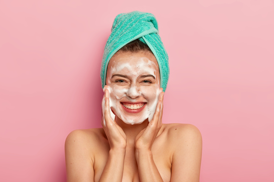 Pampering and hygiene concept. Happy young European woman massages cheeks, aplies bubble foam, washes face, smiles positively, has naked body, enjoys taking shower, wants to have clean skin.