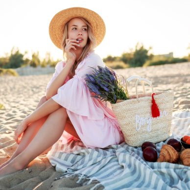 Picnic  in countryside near ocean . Graceful young woman with blond wavy hairs in elegant  pink dress  enjoying holidays and eating fruits.