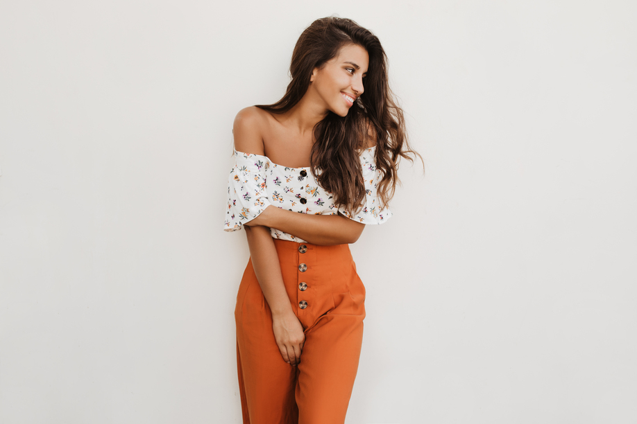 Picture of lady in orange trousers with high waist and white top with floral print. Brunette girl is cute smiling on white background