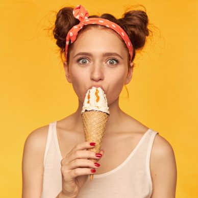 Portrait of attractive, lovely girl with two buns. Wearing white tank top and red doted hairband. Eat ice cream, watching at you with big eyes. Watching at the camera isolated over yellow background