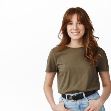 Portrait of smiling redhead girl with bangs, looking cute and happy at camera, casually standing, relaxed pose with hands in jeans pockets, white background.