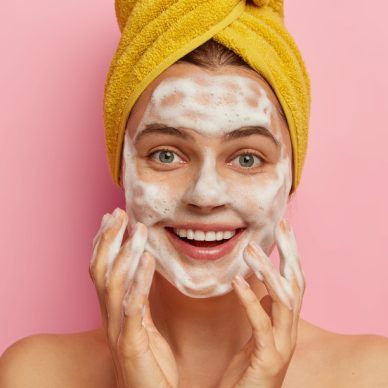 Isolated shot of pretty smiling Caucasian woman pampers face, washes skin with foaming gel, wears wrapped yellow towel on head, cares about body, isolated over pink background. Cleansing concept