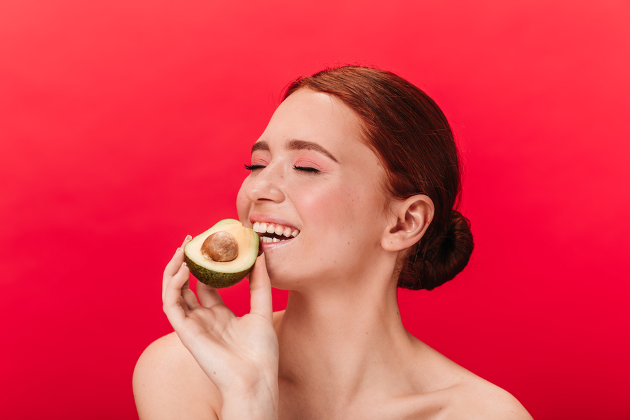 Refined girl eating avocado. Studio shot of enchanting caucasian woman posing on red background.