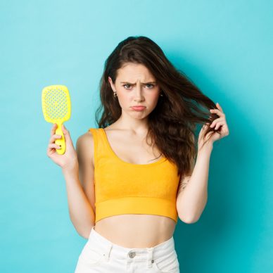 Sad young woman showing brush with strands, having hair loss problem, need cosmetic beauty treatment, standing over blue background upset.