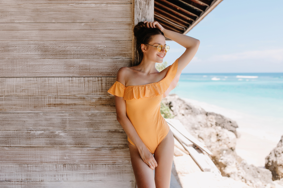 Shapely cheerful girl in orange attire posing on sea background with inspired face. Outdoor photo of spectacular young woman in sunglasses standing beside wooden house at beach.