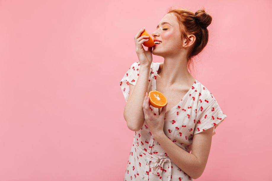 Snapshot of charming girl dressed in dress with cherry print. Woman with yellow eyeshadows holds oranges and looks into camera