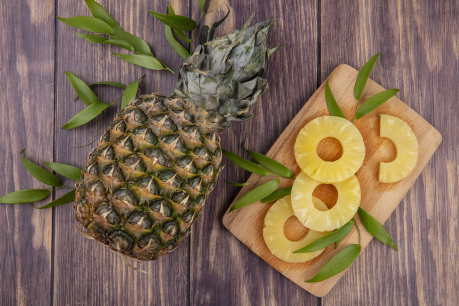 top view of pineapple and pineapple slices with leaves on cutting board and wooden background