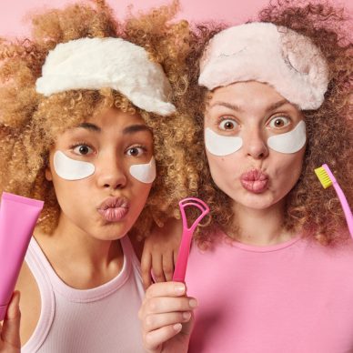 Two surprised female sisters with curly hair wears sleepmasks apply beauty patches under eyes going to clean teeth and tongue with brush stand closely to each other isolated over pink background