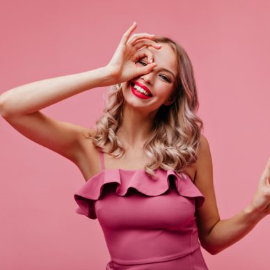 Winsome girl with blonde wavy hair fooling around on rosy background. Indoor shot of appealing caucasian woman in pink dress