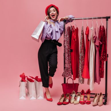 Woman in black trousers and purple blouse laughs, leaning on stand with elegant clothes on pink background