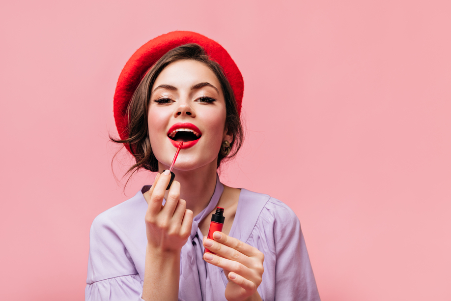 Woman in bright beret paints her lips with red lipstick. Girl in lilac blouse posing on pink background