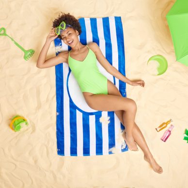 Positive Afro American woman wears green bikini and snorkeling glasses lies on blue striped towel enjoys summer vacation spends free time at sandy beach sunbathes during sunny day. Holidays.