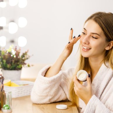 Young beautiful blonde girl cares for face skin with moisturizer in front of mirror.