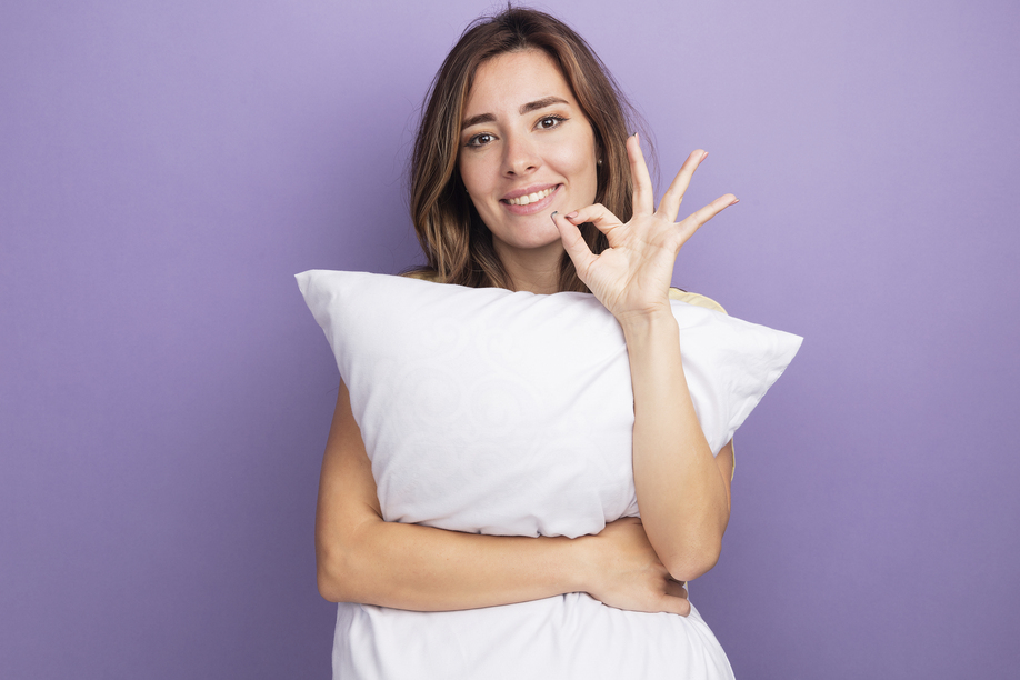 young beautiful woman in beige t-shirt holding white pillow looking at camera with smile on face doing ok sign standing over purple background