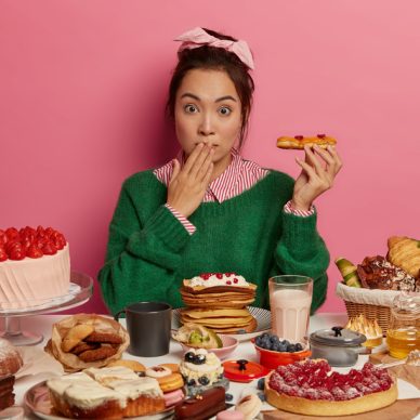 Oops, so much calories. Surprised Korean woman holds tasty pastry, eats cheat meal containing much sugar, has imbalanced nutrition and diet breakdown, poses near table with various desserts.